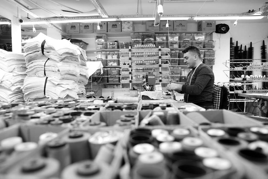 Monochrome image of a textile workshop showcasing fabric stacks and a worker at a sewing station.